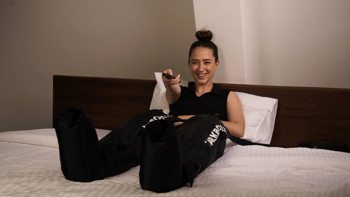 Frau liegt auf einem Bett, die Legs in den STARKBOOTS Recovery Boots, und bedient den Controller