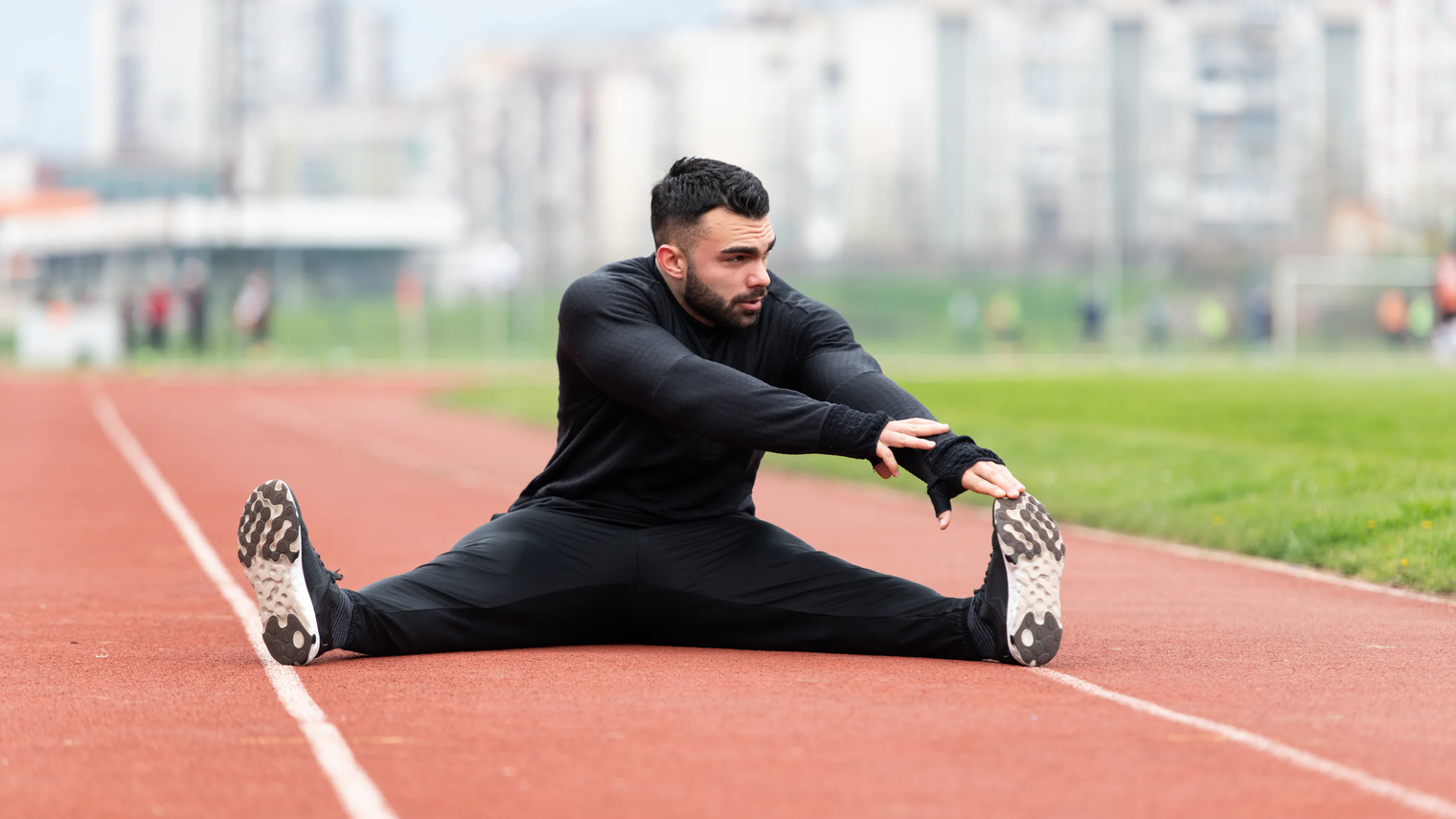 Mann in dunkler Sportkleidung dehnt sich im Sitzen auf einer roten Laufbahn