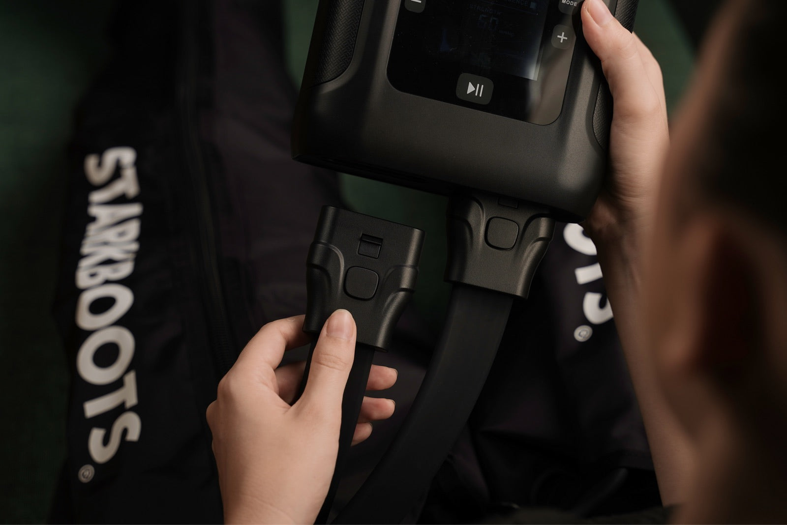 Eine Hand hält den Smart Monitor V2, die Steuereinheit der STARKBOOTS, die Boots liegen dahinter