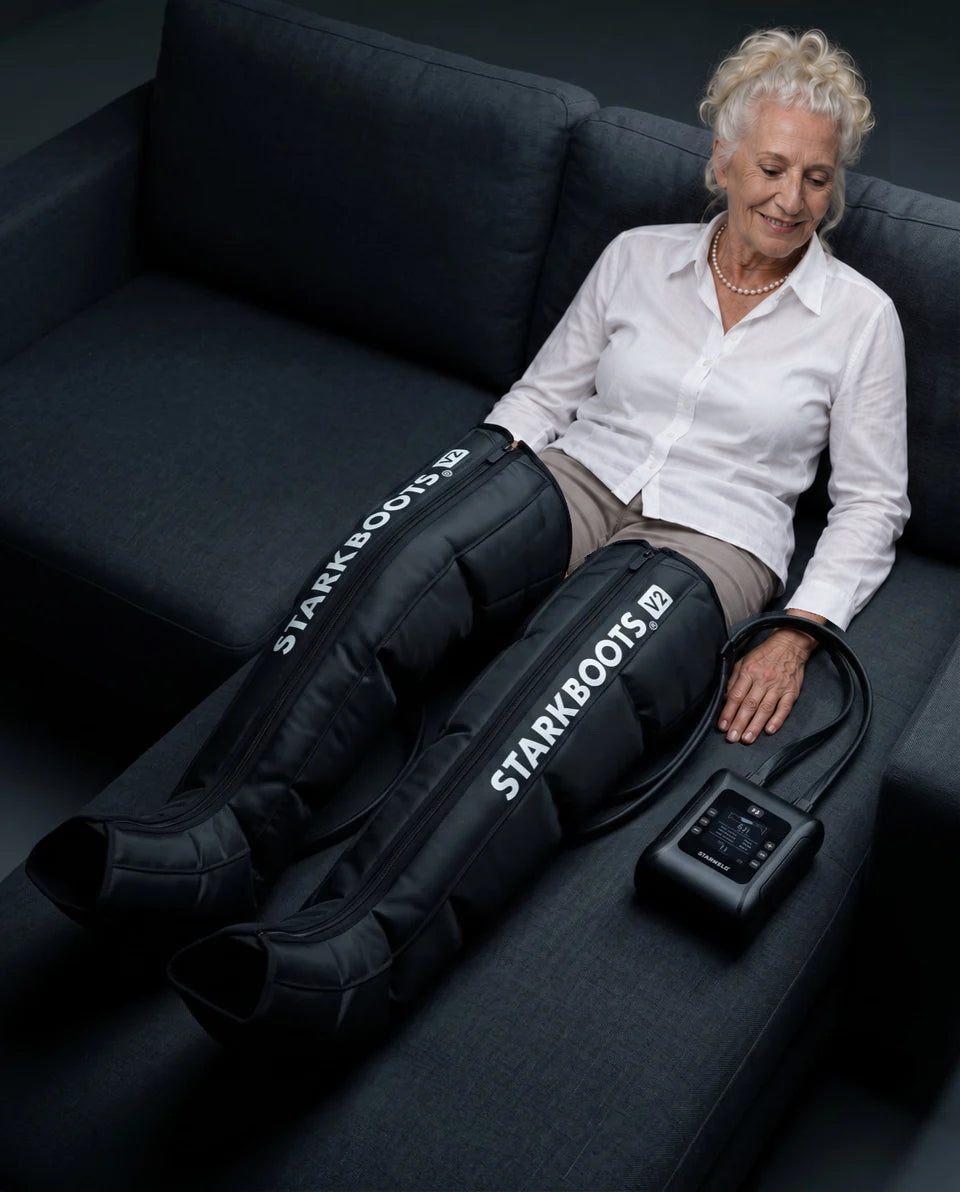 Ältere Frau sitzt mit STARKBOOTS V2 auf dem Sofa; die Steuerung liegt neben ihr.